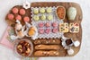 木のプレートに並べられた焼き菓子の詰め合わせ。ピンクや緑の小さな焼き菓子、フクロウ型クッキー、花柄のフロランタン、ビスコッティ、ナッツ菓子などが並び、全体を上から撮影している。