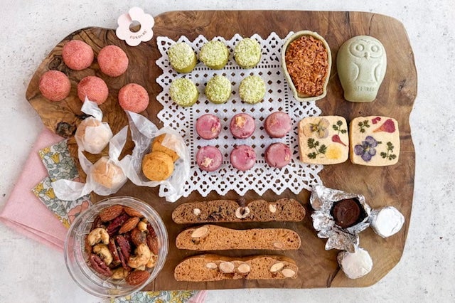 木のプレートに並べられた焼き菓子の詰め合わせ。ピンクや緑の小さな焼き菓子、フクロウ型クッキー、花柄のフロランタン、ビスコッティ、ナッツ菓子などが並び、全体を上から撮影している。