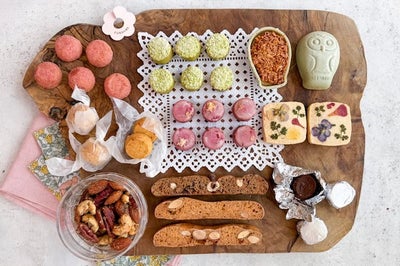 木のプレートに並べられた焼き菓子の詰め合わせ。ピンクや緑の小さな焼き菓子、フクロウ型クッキー、花柄のフロランタン、ビスコッティ、ナッツ菓子などが並び、全体を上から撮影している。