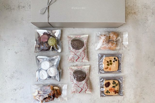 FUKU+REブランドのギフトボックスと、個包装パッケージに入ったお花の押し花クッキー、ナッツ、カラフルな焼き菓子、フクロウ型のフロランタン、アルミに包まれたラム酒のお菓子、桜島小みかんの焼菓子を並べた俯瞰写真。