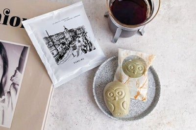 FUKU+RE(フクレ)のドリップコーヒーと、フクロウ型のフロランタンが並ぶおしゃれな朝のテーブル。コーヒーはカップに注がれており、パッケージには100年前の鹿児島の街並みが描かれている。