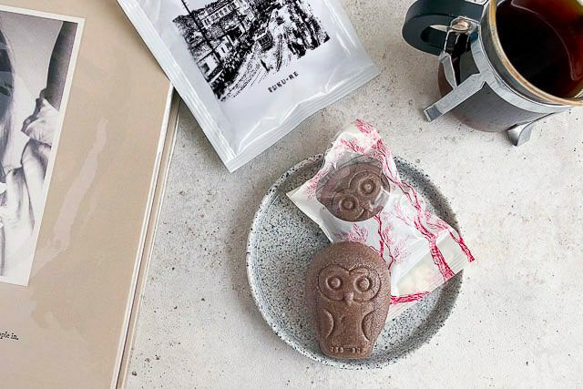 FUKU+RE(フクレ)のドリップコーヒーと、チョコレート味のフクロウ最中のフロランタンが並ぶおしゃれな朝のテーブル。コーヒーはカップに注がれており、パッケージには100年前の鹿児島の街並みが描かれている。