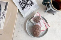 FUKU+RE(フクレ)のドリップコーヒーと、チョコレート味のフクロウ最中のフロランタンが並ぶおしゃれな朝のテーブル。コーヒーはカップに注がれており、パッケージには100年前の鹿児島の街並みが描かれている。