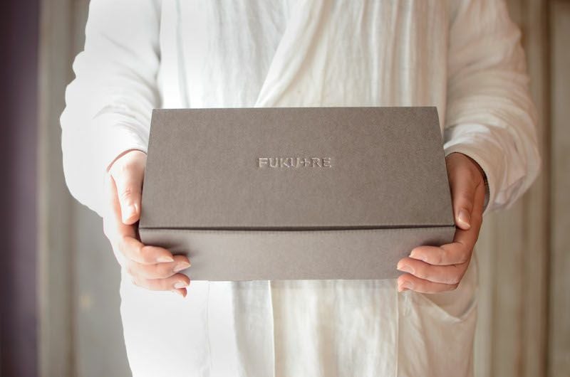 FUKU+RE(フクレ)の店員が、FUKU+REと刻印された上品なグレーのギフトボックスを両手で丁寧に持っている様子。贈り物としての丁寧さや温かみが伝わるシーン。