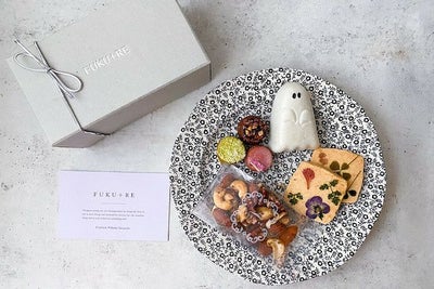 FUKU+REブランドのギフトボックスと、花の押し花クッキー、ナッツ、カラフルな焼き菓子、ゴースト型の和菓子を黒白模様の皿に盛り付けた上品なハロウィンスイーツセットの写真