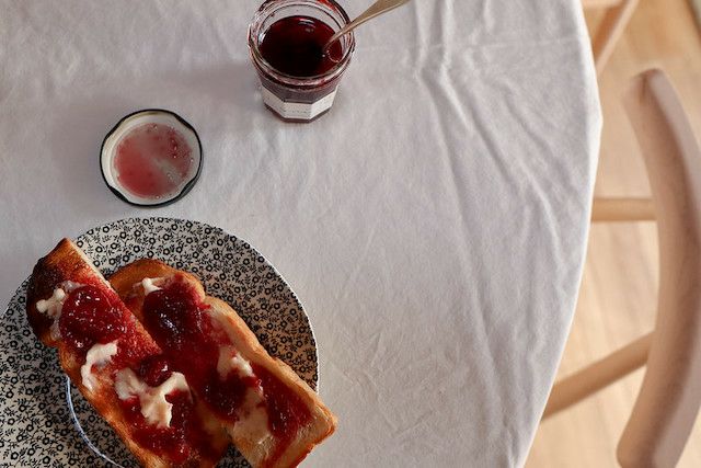 白いテーブルクロスの上で、赤いジャムを塗ったトーストと開いたジャム瓶が並ぶ。朝食の落ち着いた雰囲気が伝わるテーブル風景